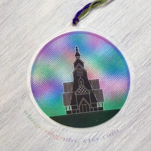 Puede incluir: Una pegatina redonda e iridiscente con una silueta negra de un edificio de iglesia. La pegatina tiene un patrón de arcoíris con tonos de rosa, morado, verde y azul. La iglesia tiene un campanario alto y un techo puntiagudo.