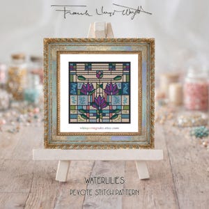 May include: A framed cross-stitch pattern of water lilies in a square, displayed on a small easel. The artwork features purple, blue, and green floral designs. The frame is gold with a distressed finish. The text "WATERLILIES DEVOTE STITCH PATTERN" is visible.