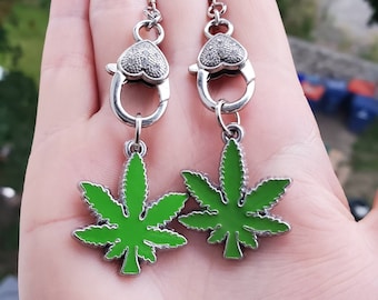 May include: A pair of silver earrings with a green marijuana leaf charm. The earrings have a heart-shaped clasp.