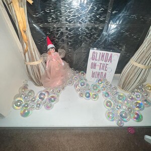 Puede incluir: Una pequeña muñeca con un vestido rosa y un gorro de elfo rojo, con un letrero que dice "Glinda on the Winda". Burbujas iridiscentes están dispuestas alrededor de la muñeca. El fondo es una ventana.