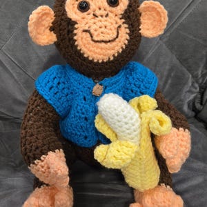 May include: A crocheted monkey toy wearing a blue sweater and holding a crocheted banana. The monkey has brown fur, a peach-colored face, and black eyes.