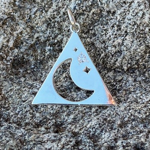 May include: A silver charm pendant shaped like a triangle with a crescent moon cut out and a small clear crystal set inside. The charm has a loop at the top for attaching to a necklace or bracelet.