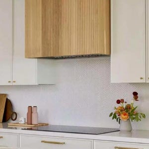 Op de afbeelding: Een moderne keuken met witte kasten, gouden handgrepen en een afzuigkap van licht hout. Een zwarte kookplaat en een vaas met bloemen voegen kleur toe. De achterwand heeft een subtiel visgraatpatroon.