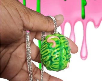 May include: A green brain pendant with pink accents hangs from a silver chain. The brain-shaped charm has detailed ridges and a realistic appearance. The background features pink and green dripping paint.