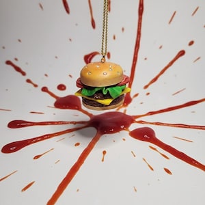 May include: A miniature hamburger pendant with a gold chain hangs against a white background splattered with red sauce. The burger features a bun, patty, cheese, lettuce, and tomato. The pendant is a unique accessory.