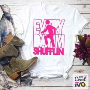 Puede incluir: Camiseta blanca con el texto "EVERY DAY I'M SHUFFLIN" en rosa brillante. El diseño incluye una figura de palo bailando. La camiseta se combina con un par de botines de ante grises y una camisa a cuadros.