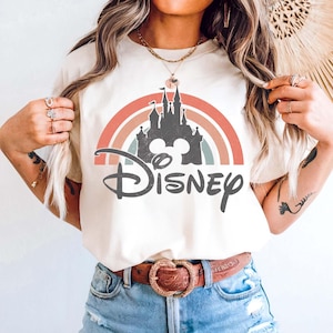May include: Ivory-colored t-shirt featuring a colorful rainbow design with a castle silhouette and the word "Disney" in a script font. The model is wearing blue denim shorts and a brown belt with a heart-shaped buckle.