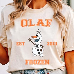 May include: A beige t-shirt featuring the word "OLAF" in orange block letters above a cartoon image of Olaf the snowman. Below the image are the words "FROZEN" and "EST 2013" in orange. The shirt is paired with denim shorts.