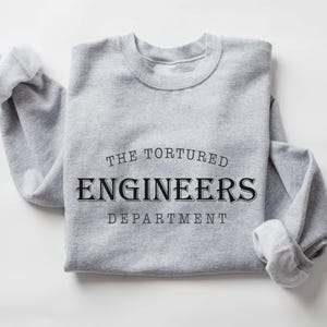 May include: A light grey sweatshirt featuring the text "THE TORTURED ENGINEERS DEPARTMENT" in black. The sleeves are rolled up. The words "sport grey" are at the bottom. The sweatshirt is folded.