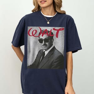 May include: A navy blue short-sleeve t-shirt featuring a black and white photographic print of a man wearing a suit and sunglasses. Above the portrait, the word WALT is printed in a bold, red, stylized script font against a light gray background.
