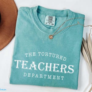 Puede incluir: Una camiseta verde agua con el texto blanco "THE TORTURED TEACHERS DEPARTMENT". Un collar dorado con un colgante descansa sobre la camiseta doblada, junto a un sombrero marr&oacute;n y una hoja de palma seca.