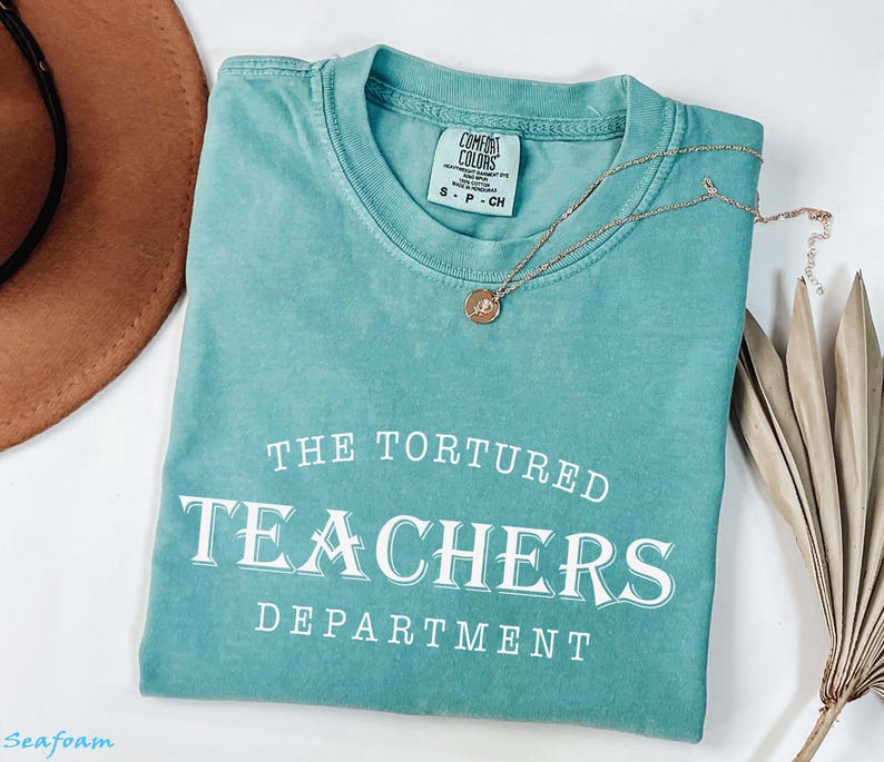 Puede incluir: Una camiseta verde agua con el texto blanco "THE TORTURED TEACHERS DEPARTMENT". Un collar dorado con un colgante descansa sobre la camiseta doblada, junto a un sombrero marr&oacute;n y una hoja de palma seca.