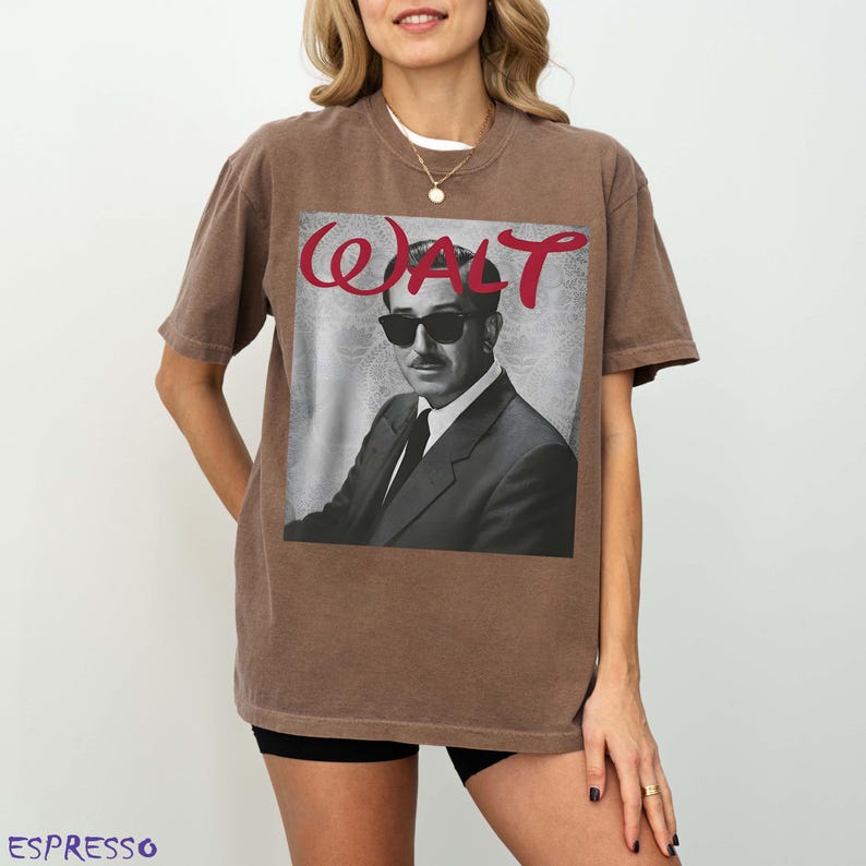 May include: A brown short-sleeve t-shirt featuring a graphic print of a man wearing sunglasses and a suit. The text "WALT" is printed in red cursive script above the portrait. The shirt has a crew neckline and a relaxed, boxy fit.