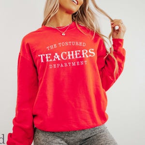 Puede incluir: Una sudadera roja con el texto "THE TORTURED TEACHERS DEPARTMENT" en letras blancas. La sudadera es de color rojo liso y tiene mangas largas. La modelo lleva pantalones grises.