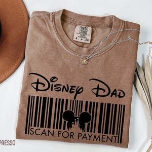 K&ouml;nnte beinhalten: Espressofarbenes T-Shirt mit dem Text "Disney Dad" &uuml;ber einem Barcode-Design mit Mickey Mouse-Ohren und den Worten "SCAN FOR PAYMENT". Eine silberne Halskette mit einem kleinen Anh&auml;nger liegt auf dem Shirt.