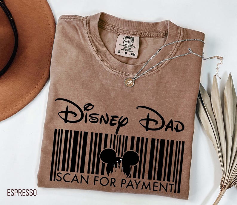 K&ouml;nnte beinhalten: Espressofarbenes T-Shirt mit dem Text "Disney Dad" &uuml;ber einem Barcode-Design mit Mickey Mouse-Ohren und den Worten "SCAN FOR PAYMENT". Eine silberne Halskette mit einem kleinen Anh&auml;nger liegt auf dem Shirt.