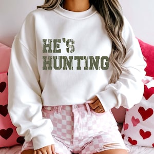 May include: A white sweatshirt with the text "HE'S HUNTING" in a distressed green font. The person is wearing pink and white checkered shorts. The background includes pink heart-shaped pillows.