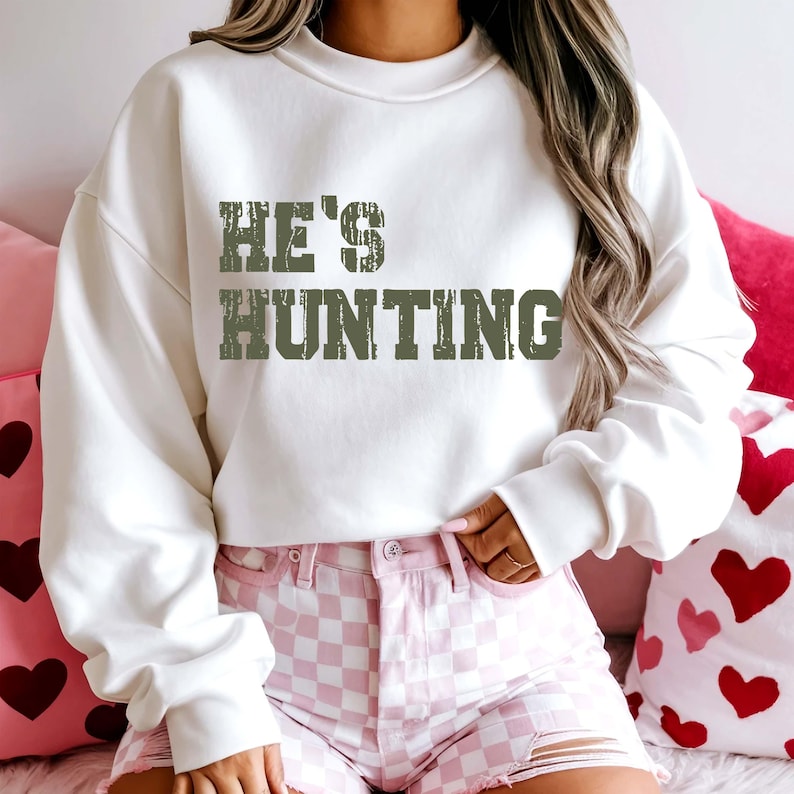 May include: A white sweatshirt with the text "HE'S HUNTING" in a distressed green font. The person is wearing pink and white checkered shorts. The background includes pink heart-shaped pillows.