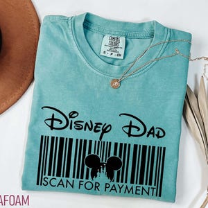 K&ouml;nnte beinhalten: T&uuml;rkisfarbenes T-Shirt mit dem Text "Disney Dad" &uuml;ber einem Barcode-Design mit Mickey Mouse-Ohren und den Worten "Scan for Payment". Eine goldene Halskette liegt auf dem Shirt.