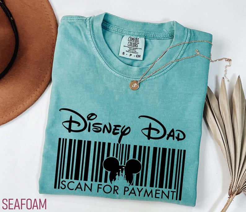 K&ouml;nnte beinhalten: T&uuml;rkisfarbenes T-Shirt mit dem Text "Disney Dad" &uuml;ber einem Barcode-Design mit Mickey Mouse-Ohren und den Worten "Scan for Payment". Eine goldene Halskette liegt auf dem Shirt.