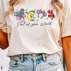 K&ouml;nnte beinhalten: Cremefarbenes T-Shirt mit einer Cartoon-Grafik und dem Text "Part of your World". Das Design umfasst einen Fisch, eine Krabbe und Micky-Maus-Ohren. Das Shirt wird mit hellblauen Jeansshorts getragen.