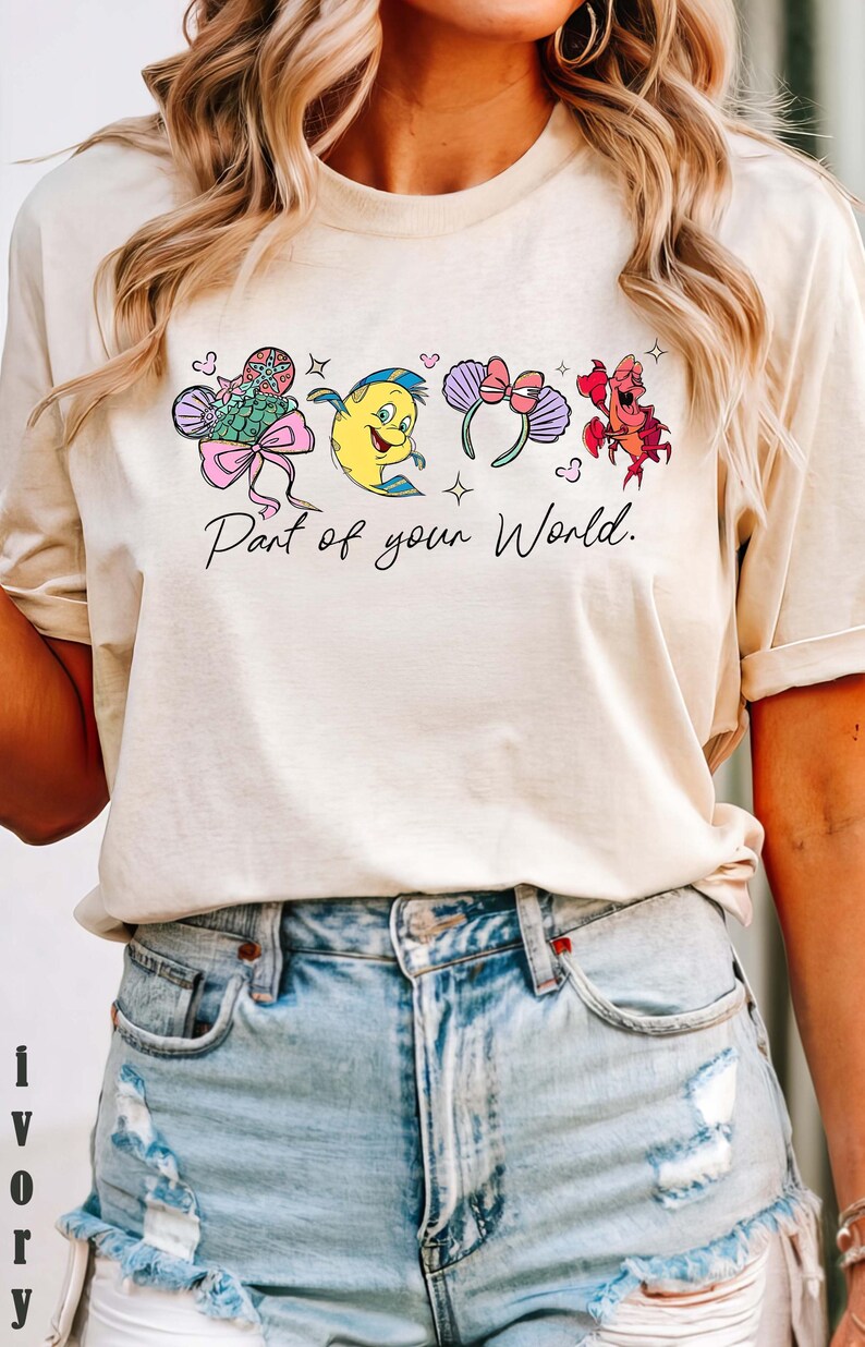 K&ouml;nnte beinhalten: Cremefarbenes T-Shirt mit einer Cartoon-Grafik und dem Text "Part of your World". Das Design umfasst einen Fisch, eine Krabbe und Micky-Maus-Ohren. Das Shirt wird mit hellblauen Jeansshorts getragen.