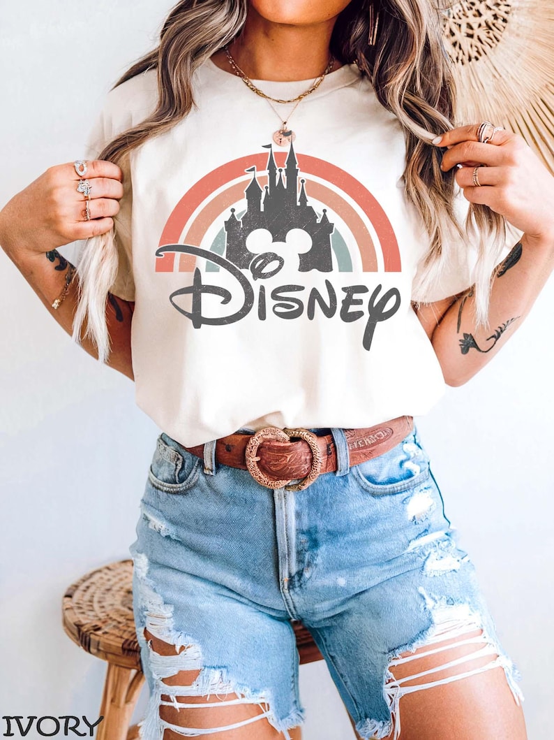 May include: Ivory-colored t-shirt featuring a colorful rainbow design with a castle silhouette and the word "Disney" in a script font. The model is wearing blue denim shorts and a brown belt with a heart-shaped buckle.