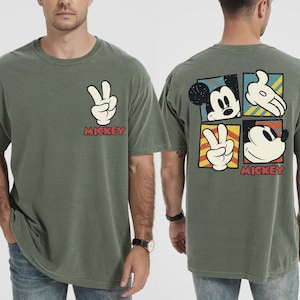 May include: A moss green t-shirt with a white peace sign and the word "Mickey" in red. The back of the shirt features Mickey Mouse imagery in a square pattern. The t-shirt is a relaxed, casual style.
