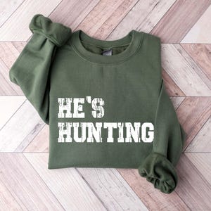 May include: An olive green sweatshirt with the text "HE'S HUNTING" in a distressed white font. The sweatshirt is folded neatly, showcasing the text prominently. The sleeves are cuffed, and the garment is made of a soft material.