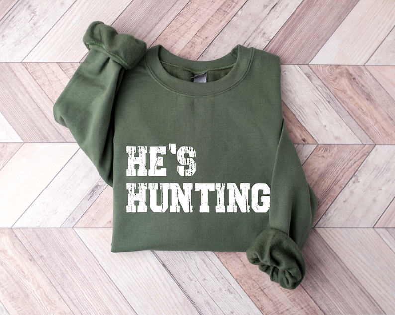 May include: An olive green sweatshirt with the text "HE'S HUNTING" in a distressed white font. The sweatshirt is folded neatly, showcasing the text prominently. The sleeves are cuffed, and the garment is made of a soft material.