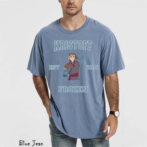 May include: A blue t-shirt featuring the word "KRISTOFF" above an illustration of the character from the movie Frozen. The shirt also includes the text "EST 2013 FROZEN". The shirt is a solid blue color.