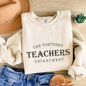 Puede incluir: Sudadera color crema con el texto negro "THE TORTURED TEACHERS DEPARTMENT". La sudadera se combina con pantalones cortos de mezclilla azules, un sombrero marr&oacute;n y sandalias. Tambi&eacute;n se ve una peque&ntilde;a planta suculenta en maceta.