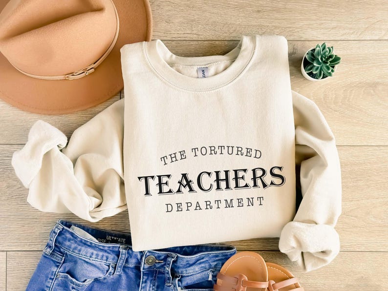 Puede incluir: Sudadera color crema con el texto negro "THE TORTURED TEACHERS DEPARTMENT". La sudadera se combina con pantalones cortos de mezclilla azules, un sombrero marr&oacute;n y sandalias. Tambi&eacute;n se ve una peque&ntilde;a planta suculenta en maceta.