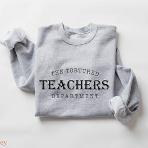 Puede incluir: Una sudadera gris claro con el texto "THE TORTURED TEACHERS DEPARTMENT" en negro. Las mangas est&aacute;n dobladas. Las palabras "sport grey" est&aacute;n en la parte inferior de la imagen. La sudadera est&aacute; doblada.