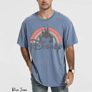 May include: Blue short-sleeved t-shirt with a Disney castle and rainbow graphic. The word "Disney" is printed below the graphic in a distressed font. The shirt is a solid blue color.