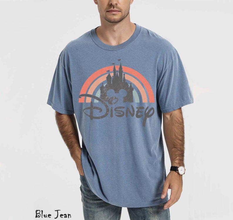 May include: Blue short-sleeved t-shirt with a Disney castle and rainbow graphic. The word "Disney" is printed below the graphic in a distressed font. The shirt is a solid blue color.