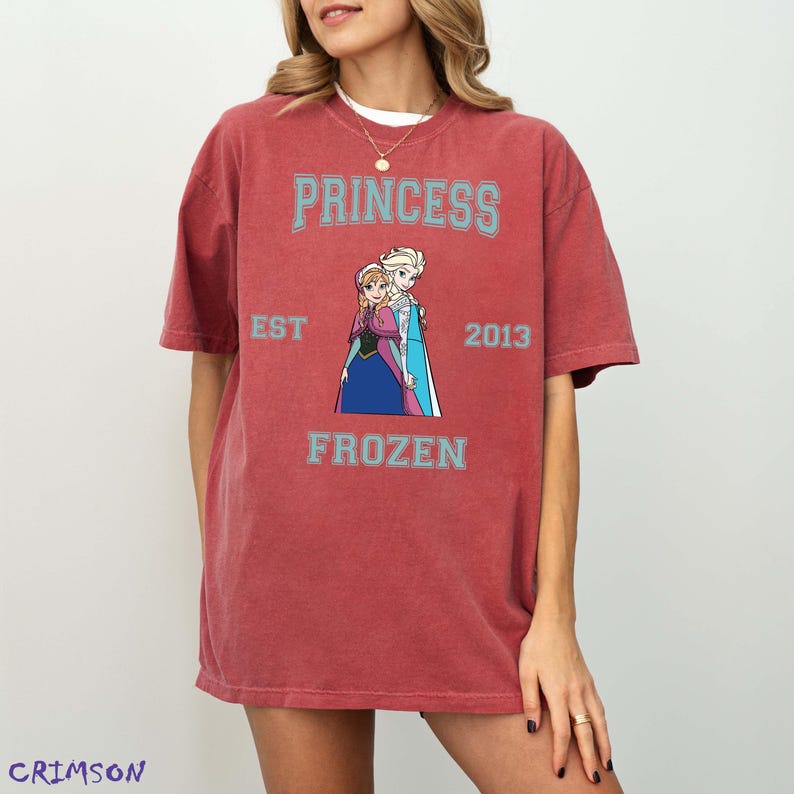 May include: A red t-shirt featuring the text "PRINCESS" above an image of two animated princesses. Below the image, the text "FROZEN" is displayed. The shirt also includes the text "EST 2013".