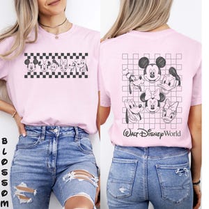 May include: A light pink t-shirt featuring black line art of Disney characters including Mickey Mouse, Minnie Mouse, Donald Duck, Daisy Duck, Goofy, and Pluto. The front displays a horizontal checkered pattern, while the back shows a larger grid with the text Walt Disney World.