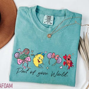 K&ouml;nnte beinhalten: T&uuml;rkisgr&uuml;nes T-Shirt mit einer Cartoon-Grafik und dem Text "Part of your World". Das Shirt hat einen Rundhalsausschnitt und kurze &Auml;rmel. Eine goldene Halskette mit Anh&auml;nger ist auf dem Shirt drapiert.