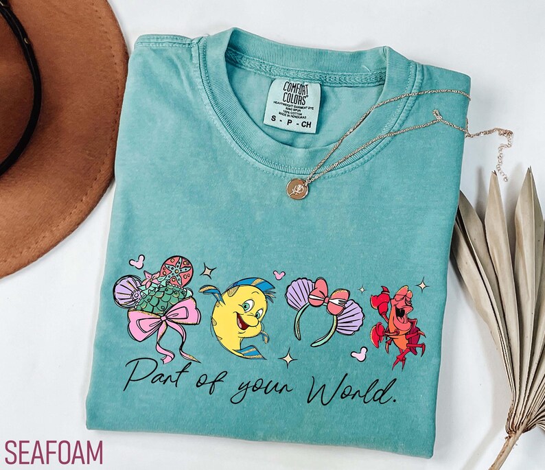 K&ouml;nnte beinhalten: T&uuml;rkisgr&uuml;nes T-Shirt mit einer Cartoon-Grafik und dem Text "Part of your World". Das Shirt hat einen Rundhalsausschnitt und kurze &Auml;rmel. Eine goldene Halskette mit Anh&auml;nger ist auf dem Shirt drapiert.