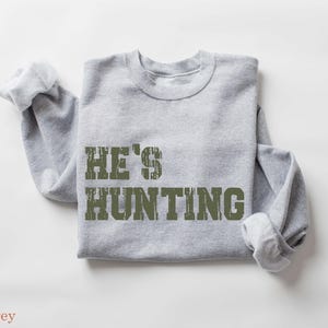 May include: A light gray sweatshirt with the text "HE'S HUNTING" in distressed olive green lettering. The long sleeves are rolled up, and the garment is displayed on a white surface. The sweatshirt is a casual apparel item.