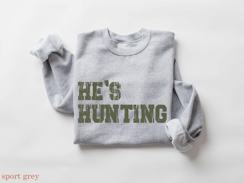 May include: A light gray sweatshirt with the text "HE'S HUNTING" in distressed olive green lettering. The long sleeves are rolled up, and the garment is displayed on a white surface. The sweatshirt is a casual apparel item.