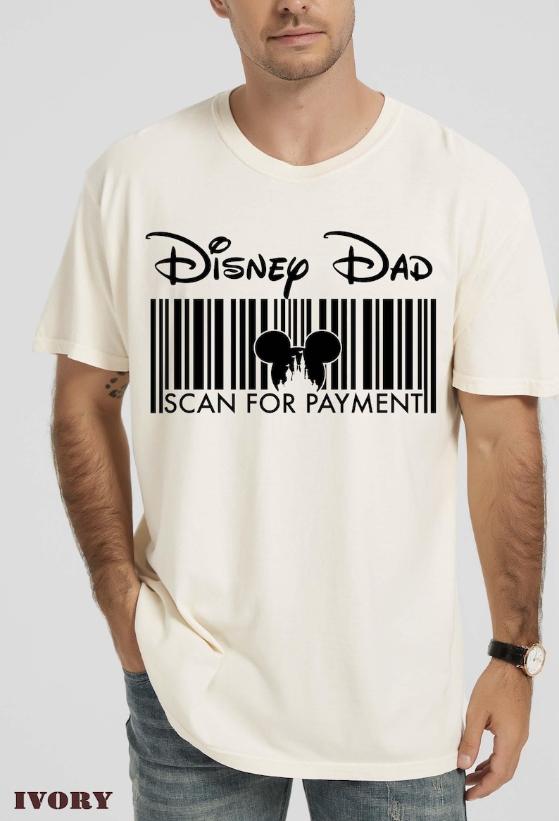 K&ouml;nnte beinhalten: Elfenbeinfarbenes T-Shirt mit dem Text "Disney Dad" &uuml;ber einem Barcode-Design. Der Barcode enth&auml;lt eine Silhouette von Mickey Mouse Ohren und einem Schloss, mit "SCAN FOR PAYMENT" darunter. Das Shirt hat kurze &Auml;rmel.