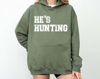 May include: Olive green hooded sweatshirt with the text "HE'S HUNTING" in white block letters. The sweatshirt has a front pocket and a drawstring hood. The model is wearing black shorts.