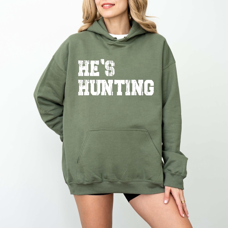 May include: Olive green hooded sweatshirt with the text "HE'S HUNTING" in white block letters. The sweatshirt has a front pocket and a drawstring hood. The model is wearing black shorts.