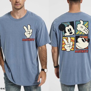 May include: Blue t-shirt with Mickey Mouse graphics. The front has a peace sign hand gesture and the word "Mickey" in red. The back features Mickey's face and hands in a square design. A casual, comfortable tee.