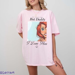 Può includere: Una maglietta rosa chiaro con un'illustrazione a cartoni animati di un personaggio con i capelli rossi e la scritta "But Daddy I Love Him". La maglietta ha una vestibilità ampia e maniche corte. Il testo "EST 1989" è sopra le parole "But Daddy".