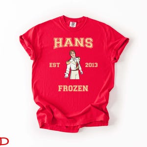 May include: A red t-shirt with the name "HANS" in gold lettering above an illustration of a character in a white and gold uniform. The text "EST 2013" and "FROZEN" are also printed on the shirt.
