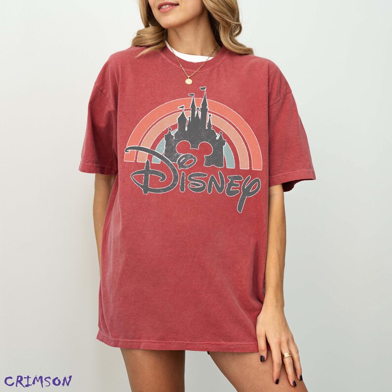 May include: A red t-shirt featuring a Disney castle design with a rainbow arch and the Disney logo. The shirt has short sleeves and a relaxed fit. The word "Crimson" is printed in the bottom left corner.