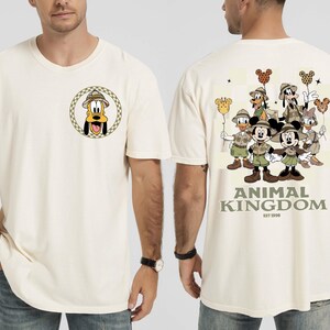 May include: Ivory t-shirt with a Pluto graphic on the front, wearing a safari hat. The back displays Disney characters like Mickey and Minnie Mouse, Donald Duck, and Goofy. The text "ANIMAL KINGDOM EST 1998" is printed below the characters.